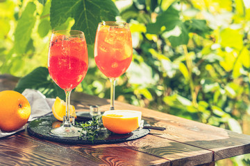 Two Aperol spritz cocktail in big wine glass with oranges, summer Italian fresh alcohol cold drink. Sunny garden with vineyard background.