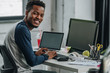 cheerful african american programmer looking at camera while sitting near computers
