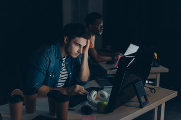 selective focus of thoughtful programmer working at night in office near african american colleague