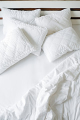 Unmade hotel room bed with bunch of puffy pillows in pile, perfectly clean snow white sheets and blanket in natural sun light. Close up, copy space for text.