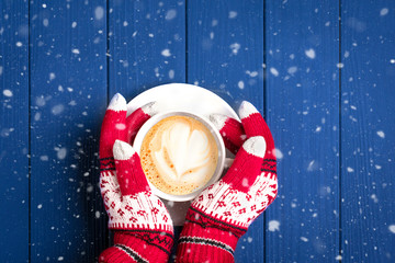 female hands with knitted gloves with New Year's pattern hold white cup with hot coffee cappuccino on blue wooden background Top view Flat lay Christmas morning concept Holiday card