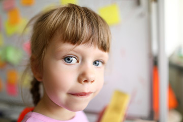 Focus on female child looking at camera with strange and unreadable glance on cute face and standing in modern classroom with high-tech glass board. Blurred background