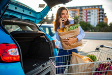 Beautiful young woman shopping in a grocery store/supermarket, putting the groceries into her car in the parking lot, looking around and talking with smartphone.
