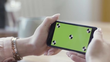 Poster - Woman looking at horizontal smartphone with green screen. Stock footage. Close up of beautiful female hands holding mobile phone with chromakey on blurred room background.