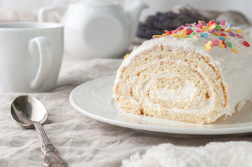 A piece of homemade biscuit roll with butter cream. Decorated with colorful topping. On a white plate. In the background is a white teapot and a mug. Close-up.