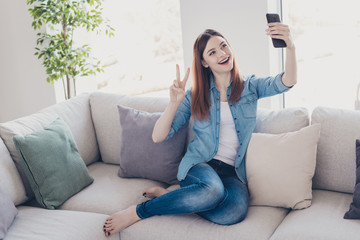 Wall Mural - Photo of pretty lady with telephone in hands making selfies showing v-sign symbol sitting comfy on sofa wearing jeans clothes apartment indoors