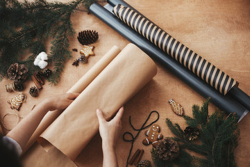 Wall Mural - Hands wrapping christmas gift in craft paper and pine branches, cones, gingerbread cookies, thread, cinnamon on rural wooden table. Wrapping stylish christmas gifts concept