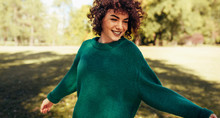 Horizontal Image Of Beautiful Young Woman Smiling Posing Against Nature Background With Windy Curly Hair, Have Positive Expression, Wearing In Green Sweater. People, Travel And Lifestyle Concept.
