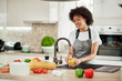 © nenadaksic - Charming mixed race woman in apron standing in kitchen ad washing yellow paprika in sink. On kitchen counter are different sorts of vegetables.