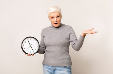 Shocked elderly woman holding wall clock and gesturing with hand