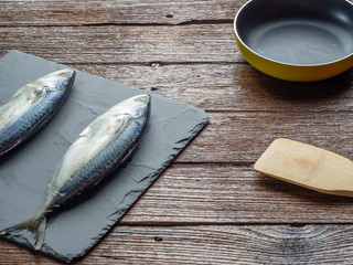 2 raw mackerel on stone cutting board with kitchen equipment on wood table.