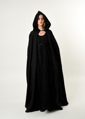 Wall Mural - full length portrait of blonde girl wearing long black flowing cloak, standing pose  with  a white studio background.