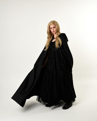 Wall Mural - full length portrait of blonde girl wearing long black flowing cloak, standing pose  with  a white studio background.