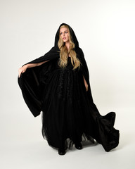 Wall Mural - full length portrait of blonde girl wearing long black flowing cloak, standing pose  with  a white studio background.