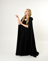 Wall Mural - full length portrait of blonde girl wearing long black flowing cloak, standing pose  with  a white studio background.