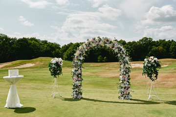 Beautiful wedding arch decorated with white and pink rose flowers on green golf course outdoors, copy space. Wedding setup. Place for ceremony. Floral composition