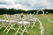 © mirage_studio - White wooden chairs with rose flowers on each side of archway outdoors, copy space. Empty chairs for guests prepared for wedding ceremony on golf course