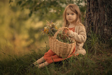 Small Girl Sitting Under The Tree With Wicker Basket Full Of Wildflowers And Looking At The Camera