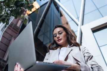 Close-up of young professional businesswoman using her laptop outside while working at modern business center, modern buildings in the background