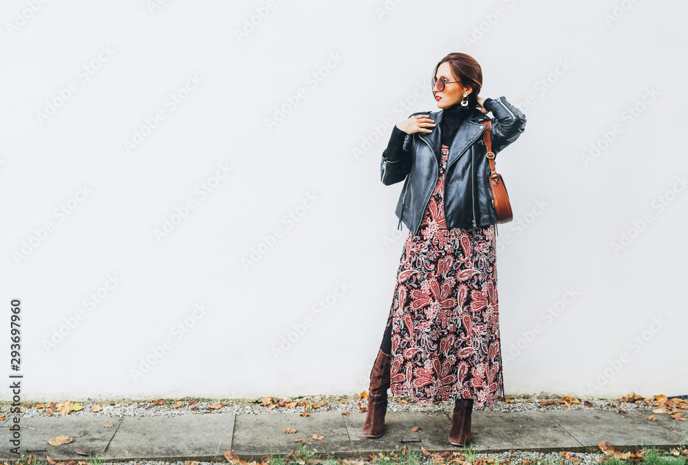 Obraz na płótnie Smiling female dressed boho fashion style colorful long dress with black leather biker jacket with brown leather flap bag posing on the white wall background. w salonie