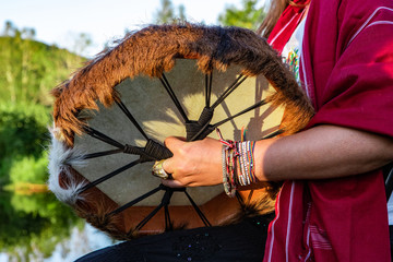 Wall Mural - Sacred drums during spiritual singing. A spiritual music player is seen close up, wearing traditional bracelets and clothing as he holds a Native American drum during a music celebration in a park.