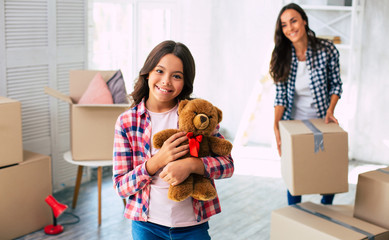 Wall Mural - Look what I have! Close-up photo of a lovely little girl smiling and holding a teddy bear in a studio while her mother moves cardboard boxes and smiles on the background.