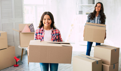 New life waits ahead. Let me help you! Small kid is smiling while helping her young beautiful mother with cardboard boxes relocation in their new home.