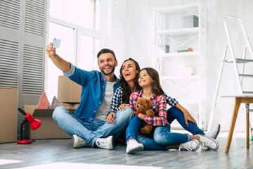 Wall Mural - Like in a fairy tale. Young family of father, mother and daughter are sitting on the floor in the a new apartment laughing and taking a selfie together.