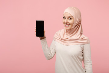 Smiling young arabian muslim woman in hijab light clothes posing isolated on pink background. People religious Islam lifestyle concept. Mock up copy space. Hold mobile phone with blank empty screen.