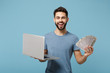 © ViDi Studio - Young smiling joyful man in casual clothes posing isolated on blue wall background. People lifestyle concept. Mock up copy space. Holding laptop pc computer, fan of cash money in dollar banknotes.