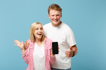 Wall Mural - Funny couple two friends guy girl in white pink t-shirts posing isolated on blue background. People lifestyle concept. Mock up copy space. Hold mobile phone with empty blank screen, showing thumb up.