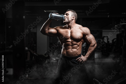 Muscular athletic bodybuilder fitness model posing and drinks water after exercises in gym