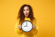 Amazed Young African American Girl In Fur Sweater Posing Isolated On Yellow Orange Background, Studio Portrait. People Lifestyle Concept. Mock Up Copy Space. Holding Clock, Keeping Mouth Wide Open.