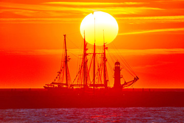  A sailing ship of the regatta 'Hanse Sail' for traditional sailing ships at sunset at the lighthouse of Rostock-Warnemünde.