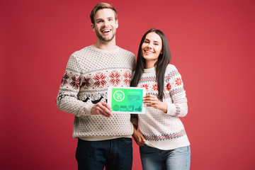 smiling couple in winter sweaters showing digital tablet with shopping app isolated on red