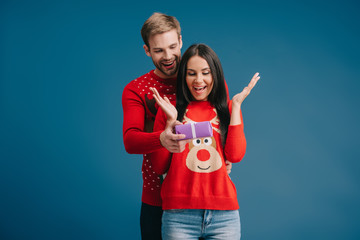 smiling man presenting christmas gift to surprised woman isolated on blue