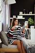 © chika_milan - Beautiful smiling caucasian brunette dressed in sweater sitting on bed in bedroom, using tablet and drinking coffee in morning.