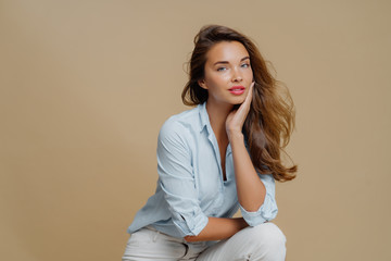 Portrait of attractive young woman sits against brown background, touches cheek, dressed in fashionbale outfit, has thoughtful look at camera, demonstrates her beauty. People and style concept