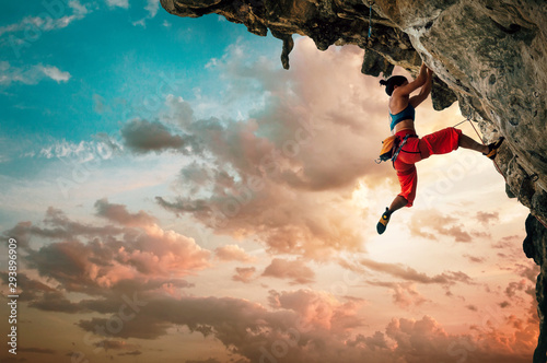 Fotomural Athletic Woman climbing on overhanging cliff rock with sunset sky background
