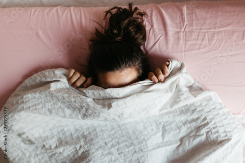 Fotomural Playful young woman hiding face under blanket while lying in cozy bed on white pillow, pretty curious girl feeling shy peeking from duvet, covering with white sheet, head shot close up