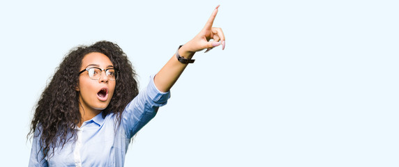 Canvas Print - Young beautiful business girl with curly hair wearing glasses Pointing with finger surprised ahead, open mouth amazed expression, something in front