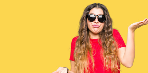 Poster - Young beautiful woman wearing red t-shirt and sunglasses clueless and confused expression with arms and hands raised. Doubt concept.