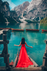 Lady in red long dress on the background of the turquoise lake with wooden boats in mountains. Brunette woman back view like a fairytale princess. Dolomites Alps, lago di Braies, Italy