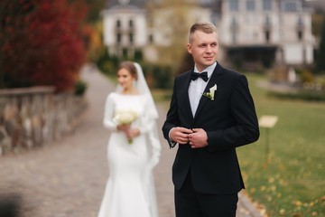 Stylish couple walking in the park at their wedding day. Happy newlyweds outside in autumn weather. Groom staind in front of bride