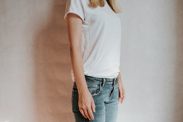 Wall Mural - Blond hair woman posing near a light wall. Beautiful young caucasian girl. Hand gestures. Emotion. Casual clothing. Studio model in work. Strong woman, future is female. Jeans and white blank t-shirt