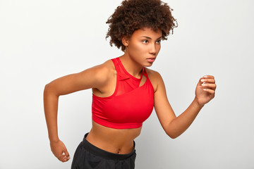 Athletic self confident woman stands in running pose, looks seriously on finish, demonstrates endurance, wears red top and black shorts, moves actively with arms, isolated on white background