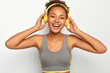 © WHstudio Leushin N - Sporty lifestyle and leisure concept. Overjoyed Afro woman smiles broadly, wears yellow headphones, listens music during fitness, uses special app, dressed in grey top, isolated over white background.