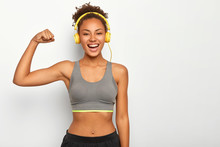 Horizontal View Of Dark Skinned Woman In Good Mood, Raises Arm With Muscles, Has Strong Body, Dressed In Gym Outfit, Listens Audio Via Modern Headphones, Poses Indoor. Fitness And Music Concept