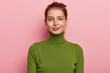 Portrait of beautiful young lady has dark combed hair, appealing appearance, wears casual green turtleneck, looks gladfully into camera, poses against pink background, feels pleased to have day off