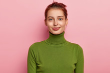 Portrait Of Beautiful Young Lady Has Dark Combed Hair, Appealing Appearance, Wears Casual Green Turtleneck, Looks Gladfully Into Camera, Poses Against Pink Background, Feels Pleased To Have Day Off